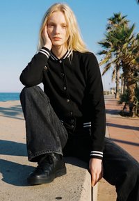 Young woman with long blonde hair, black varsity jacket, and wide black jeans sitting thoughtfully on a seaside concrete ledge near palm trees.