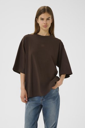 Young woman with long brown hair wears oversized dark brown T-shirt and blue jeans, standing against plain light background.