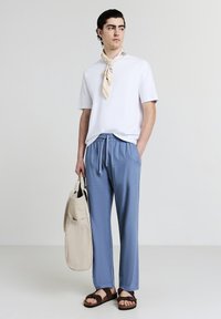 Weißes kurzärmliges Hemd, hellblaue Hose mit Kordelzug, braune Sandalen und eine beigefarbene Tasche. Seidenschal um den Hals gebunden, glattes Material und lockere Passform.
