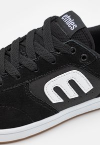 Μαύρα σουέτ αθλητικά παπούτσια με λευκό δερμάτινο λογότυπο. Διαθέτουν μαύρες κορδόνες, επενδυμένο κολάρο και ελαστική σόλα. Μινιμαλιστικός σχεδιασμός με καθαρές γραμμές.