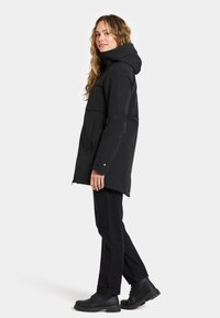 Black waterproof jacket with a hood, front pockets, and a zip closure. Worn with black pants and boots, featuring a straight cut design.