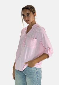 Jasnoróżowa koszula z długim rękawem z kołnierzykiem mandarynkowym, z kieszonką z przodu i podwiniętymi rękawami, w połączeniu z jasnoniebieskimi dżinsami. Teksturowany materiał.