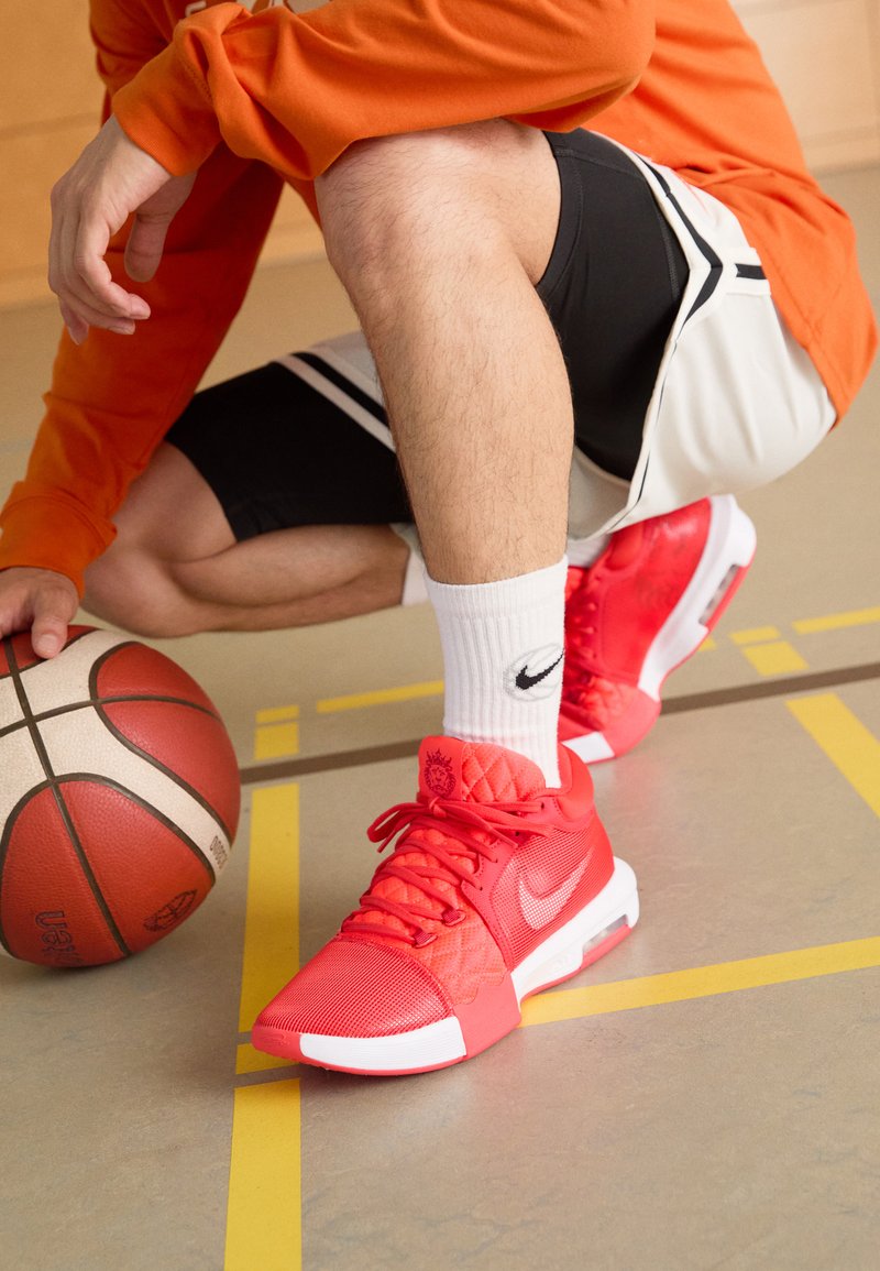 Crvene Nike košarkaške tenisice s teksturiranim mrežastim gornjim dijelom, podstavljenim ovratnikom i bijelim naglascima. Košarka i označene linije na terenu su vidljive.