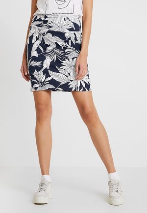 Kvinde iført en marineblå nederdel med hvidt blomsterprint, kombineret med hvide sneakers og hvide ankelsokker, står foran en ensfarvet baggrund.