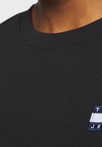 Zwarte katoenen t-shirt met een ronde hals, korte mouwen en een klein logo-patch met blauwe en witte strepen aan de linkerkant.