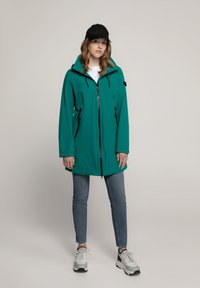 Türkisfarbene wasserdichte Jacke mit durchgehendem Reißverschluss, Kordelzugkapuze und mehreren Reißverschlusstaschen. Kombiniert mit hellblauen Jeans und grauen Sneakers.