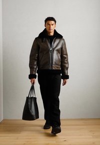Giacca bomber in pelle marrone con collo in shearling nero, chiusura con zip e polsini a costine. Il modello tiene in mano una borsa a tracolla nera, indossando pantaloni neri.