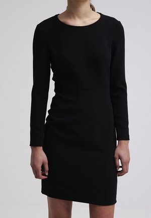 Žena v těsně přiléhajících dlouhých černých šatech s kulatým výstřihem, stojící před jednoduchým šedým pozadím.