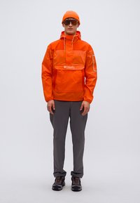 Πορτοκαλί αδιάβροχο μπουφάν τύπου πουλόβερ με μπροστινή τσέπη και κουκούλα με κορδόνι, σε συνδυασμό με γκρι παντελόνι και μαύρα παπούτσια πεζοπορίας, σε ουδέτερο φόντο.