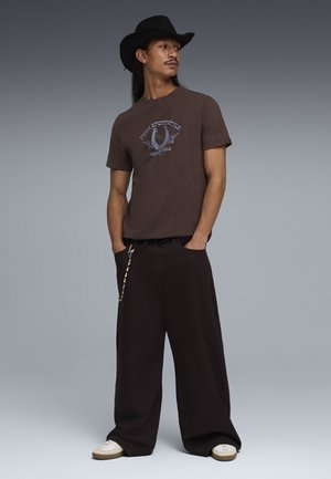 Uomo con cappello da cowboy nero, t-shirt marrone con grafica, pantaloni neri larghi con catena di perline e scarpe da ginnastica bianche, in piedi con le mani in tasca.