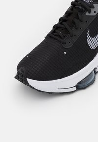 Czarny but sportowy z teksturowaną syntetyczną cholewką, białą podeszwą i widoczną jednostką powietrzną. Posiada akcenty z siateczki oraz kontrastowe logo.