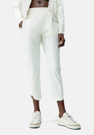 Pantalones deportivos blancos con una textura suave, silueta ajustada, bolsillos laterales y un corte entallado, combinados con zapatillas beige y blancas.