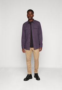 Vijolična flanelasta srajca z dvema žepoma na prsih, nošena čez črno majico. Ujemajo se z bež hlačami in črnimi čevlji na vezalke.