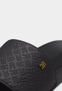 Schwarze Slidesandale mit strukturiertem Riemen, der ein geprägtes Buchstabenmuster und ein kleines goldfarbenes Metalllogo an der Seite aufweist.