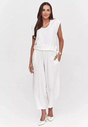 Kobieta stojąca w białym, luźnym topie bez rękawów i szerokich spodniach, na szpilkach, z ręką w kieszeni, uśmiechająca się na białym tle.
