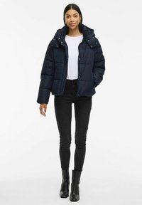 Tumed puffer jope, millel on tõmblukk, eemaldatav kapuuts ja elastsed mansetid. Kandmine valge särgi, mustade kitsaste teksade ja saapadega.