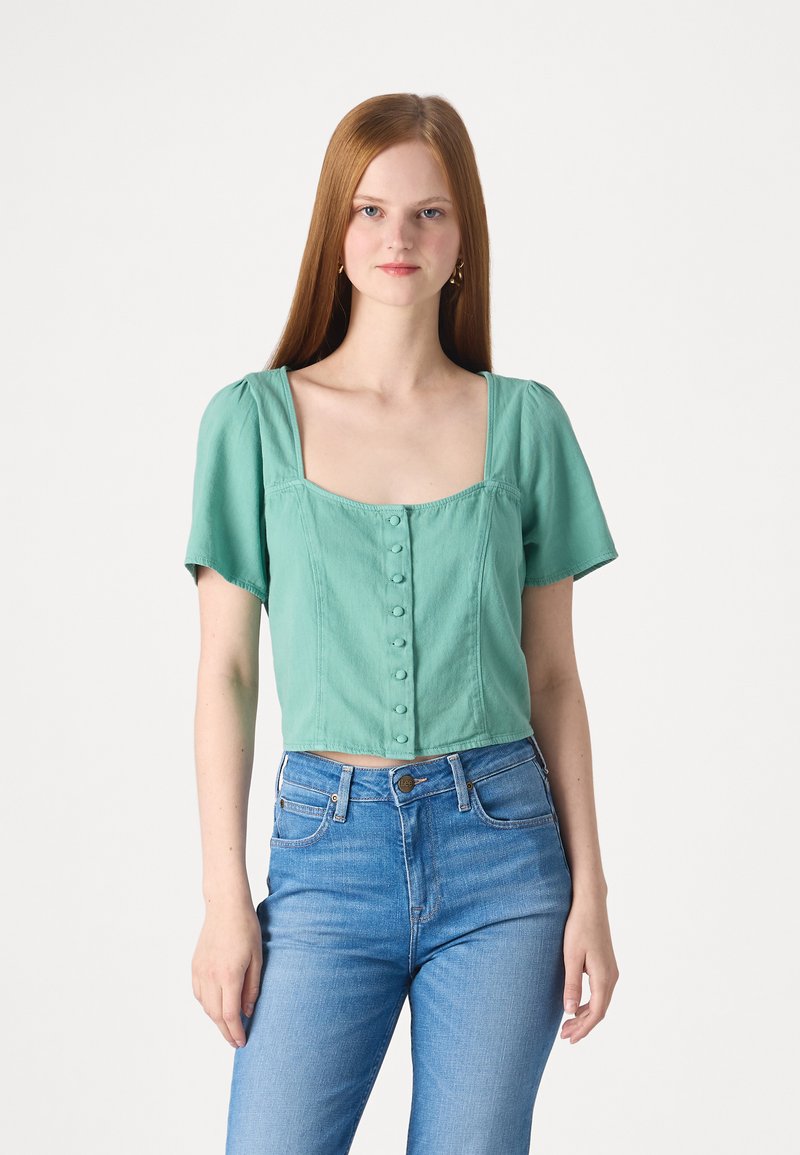 Top corto verde menta con maniche a sbuffo corte, scollo quadrato e chiusura frontale con bottoni. Abbinato a jeans di denim blu.