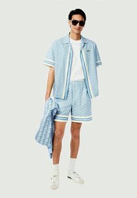 Completo composto da camicia a maniche corte azzurro chiaro e pantaloni corti con motivo geometrico, dettagli bianchi e vestibilità comoda. Il modello indossa occhiali da sole e sneakers.