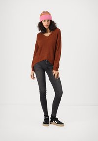 Roestkleurige, oversized gebreide trui met V-hals, gecombineerd met zwarte skinny jeans en zwarte sneakers. Een roze gestreepte beanie maakt de look af.