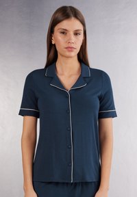 Marinblå pyjamasöverdela i mjukt material, med klassisk krage, korta ärmar och vit piping längs kanterna samt fem knappar för stängning.