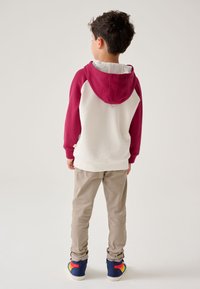 Vaikiškas kreminės spalvos megztinis su bordo spalvos rankovėmis ir gobtuvu. Yra kango kišenė ir ribuoti rankogaliai. Dėvimas su smėlio spalvos kelnėmis ir spalvingais sportbačiais.