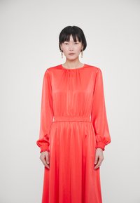 Langärmliges, bodenlanges Kleid aus lebhaftem korallfarbenem Satin mit gerafftem Ausschnitt und Rüschensaum, das an der Taille mit einem elastischen Bund für eine schöne Silhouette versehen ist.