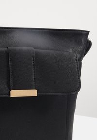 Czarna skórzana torebka o fakturowanej powierzchni, z klapką do składania i złotym akcentem na zapięciu z przodu. Prosty, prostokątny kształt.