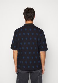 Polo a maniche corte, tessuto nero con un motivo a cuori blu e verdi ripetuto, collare piatto e vestibilità comoda.