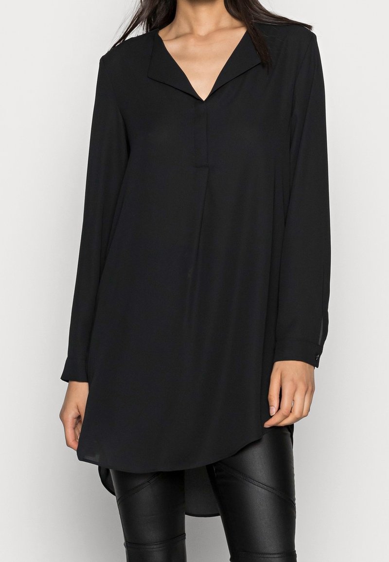 Femme portant une tunique noire à manches longues avec un col en V et un ourlet asymétrique, associée à un pantalon en cuir noir.