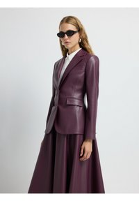Giacca in pelle bordeaux con design strutturato, rever a punta e due tasche frontali, abbinata a una lunga gonna fluida dello stesso colore.