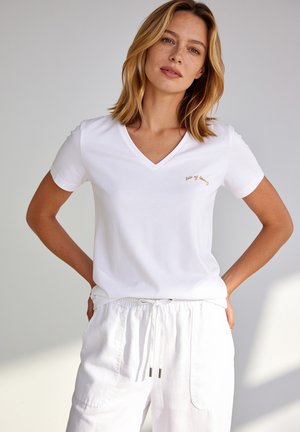 Frau mit schulterlangem blondem Haar, die ein weißes V-Ausschnitt-T-Shirt und weiße Kordelhose vor einem einfarbigen hellen Hintergrund trägt.