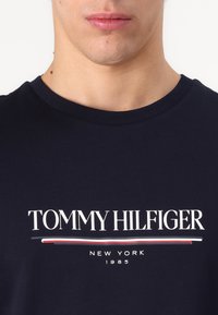 Marineblå bomulds t-shirt med hvid "TOMMY HILFIGER" logo, røde og hvide accentstriber, rund halsudskæring og korte ærmer.