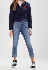 Navy pullover jacket, quarter-zip with red heart logo, paired with light blue rolled-cuff jeans and black sneakers, featuring white laces.