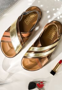 Sandales en cuir avec des brides croisées en or et bronze sur une surface blanche, à côté d'un rouge à lèvres rouge et de clés de voiture.