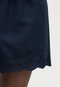 Πτερύγιο φούστας σε ναυτικό μπλε με κεντημένη λουλουδάτη διακοσμητική τρύπα, φορεμένη πάνω σε πόδι με σκούρο τόνο δέρματος σε ανοιχτόφωτο φόντο.