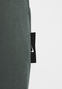 Σκούρο πράσινο βαμβακερό ύφασμα με λεία υφή; διαθέτει μια μαύρη ετικέτα με λευκό λογότυπο κοντά στη ραφή. Απλός, μινιμαλιστικός σχεδιασμός.