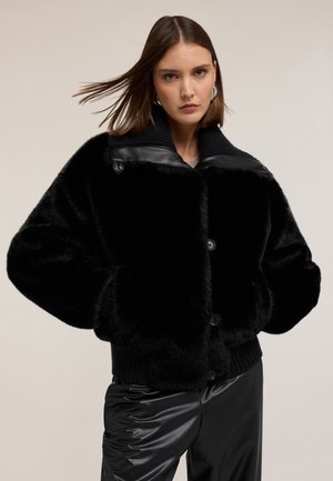 Zwarte nepbontjas met een geribbelde zoom, voorzien van een leren kraag en grote knopen. Zachte textuur met een boxy silhouet.