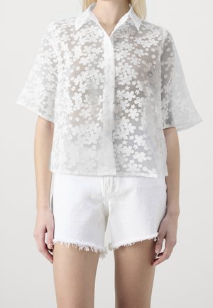 Woman wearing a white sheer floral shirt with short sleeves and white frayed denim shorts, standing against a plain background.