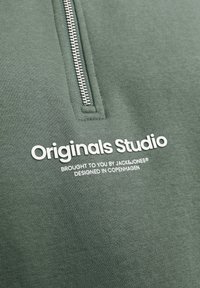 Sudadera verde con cremallera, de tela texturizada, con cremallera corta y texto impreso en blanco que detalla la marca y el origen del diseño.