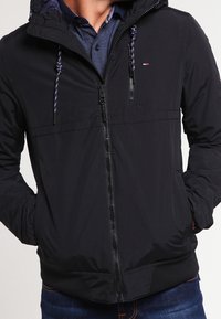 Giacca nera con cappuccio realizzata in tessuto leggero, dotata di chiusura con zip, polsini elasticizzati e dettagli con cordini. Include un piccolo logo sul petto.