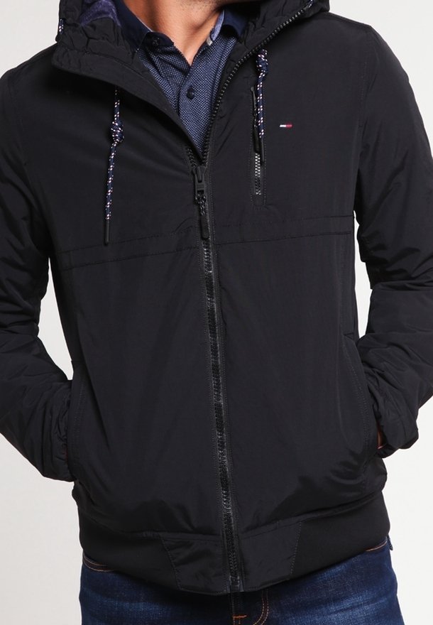 Giacca nera con cappuccio realizzata in tessuto leggero, dotata di chiusura con zip, polsini elasticizzati e dettagli con cordini. Include un piccolo logo sul petto.