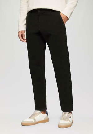 Schwarze Baumwollhosen im Slim-Fit-Stil, ausgestattet mit Fronttaschen und einem Knopfverschluss, kombiniert mit beigen und marineblauen Sneakers.