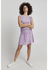 Haut lilas avec des manches volantées associé à une jupe lilas arborant un motif de feuilles blanches ; le mannequin se tient en baskets blanches, les bras croisés.