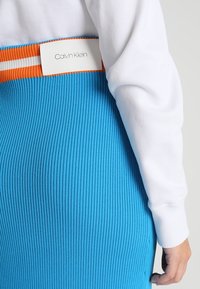 Jupe en tricot côtelé bleu avec une taille rayée orange et blanche, arborant une étiquette blanche avec "Calvin Klein". Manche blanche partiellement visible.