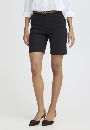 Femeie purtând pantaloni scurți negri, croiți până la genunchi, cu o curea maro, bluza albă cu mânecile surogate și pantofi cu toc închis maro, cu barete și vârf ascuțit.