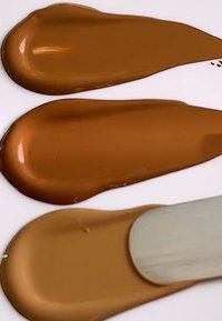 Kolme värimallia kermaisesta meikkivoiteesta eri ruskeissa sävyissä valkoisella pinnalla, joissa on sileä rakenne ja kiiltävä pinta, spatula vieressä.