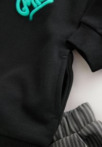 Sort sweatshirt med turkise bogstaver og ribbede manchetter, parret med grå og sorte stribede sweatpants med elastisk talje.