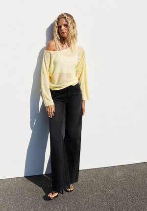 Femme blonde debout contre un mur blanc portant un haut jaune transparent à épaules dénudées, un jean noir large et des sandales noires à talons.