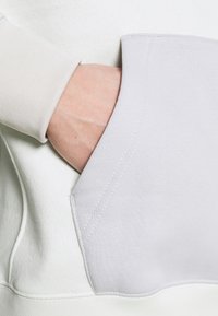 Ljusgrå sweatshirt med en känguruficka, som har ribbade ärmslut och en slät textur. Fickan har kontrasterande accentstickningar.