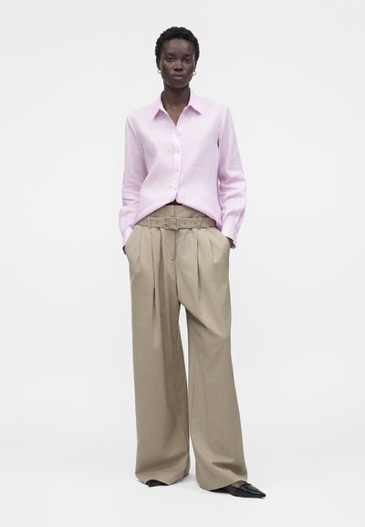 Γυναίκα φοράει ανοιχτό ροζ πουκάμισο με κουμπιά, μπουζαρισμένο μέσα σε μπεζ, ψηλόμεση φαρδιά παντελόνια με ζώνη και μαύρα μυτερά παπούτσια.